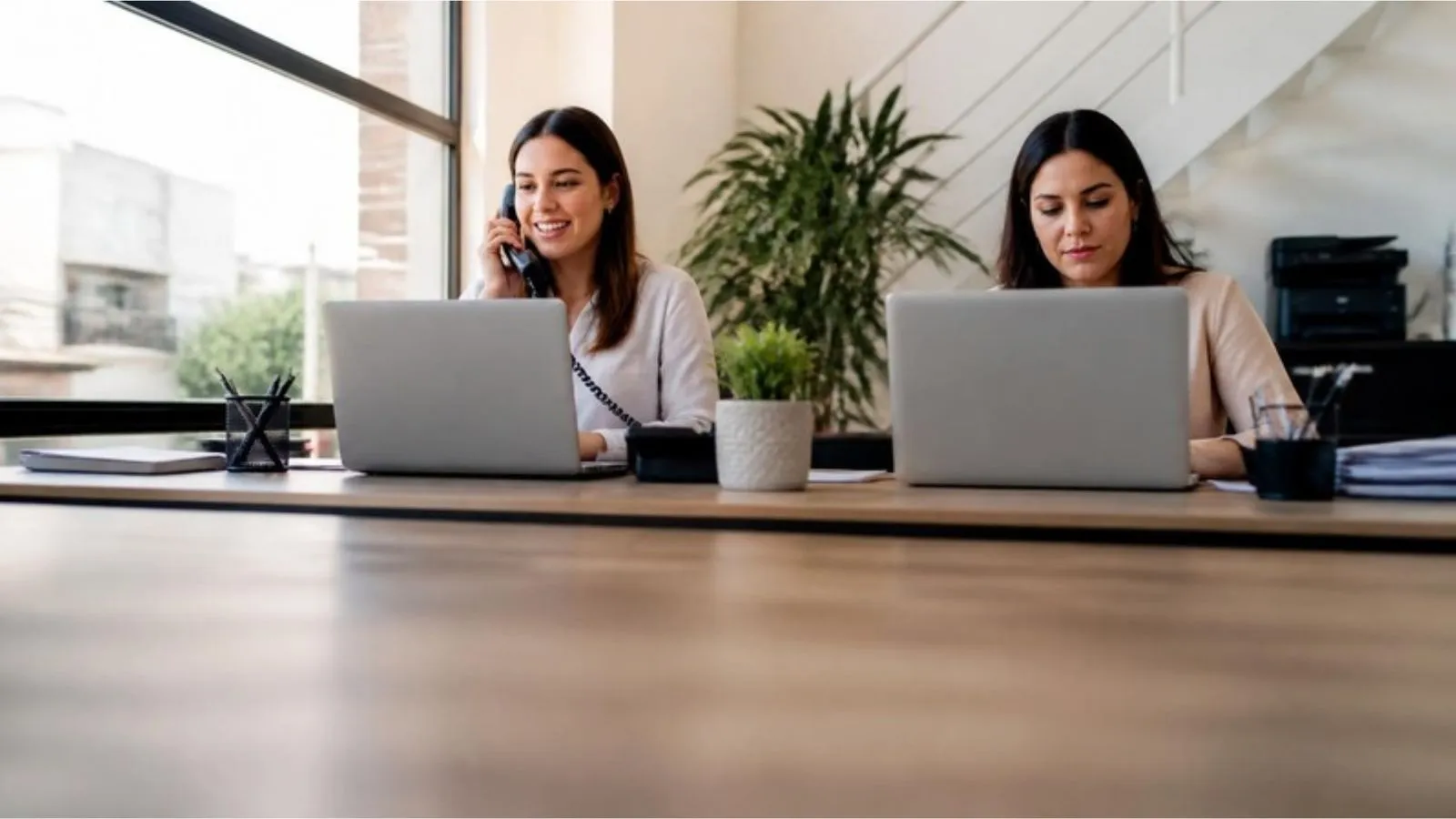 Duas contadoras trabalham em notebooks num escritório claro; uma delas fala ao telefone