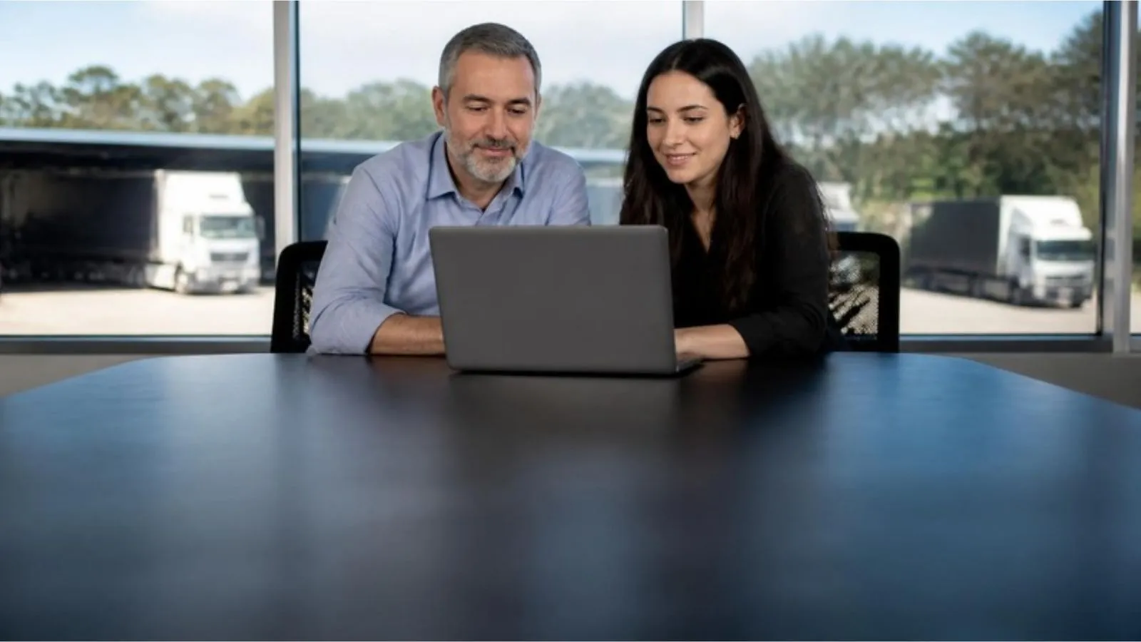 Dono de uma transportadora e uma analista de TI olham juntos um notebook na sala de reunião